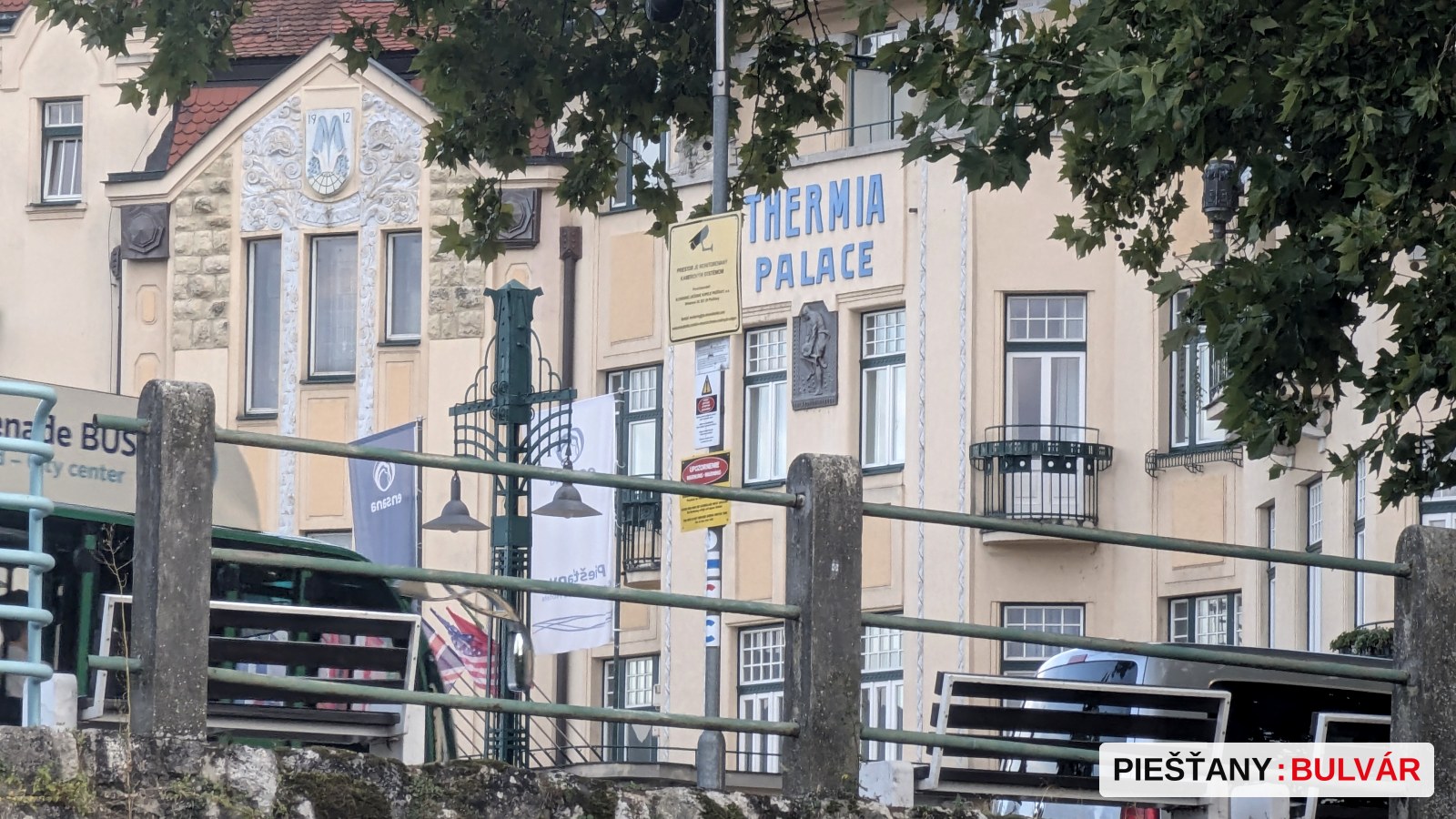 Fotografia zachytáva budovu s názvom Thermia Palace, s viditeľným názvom na fasáde a okolitou zeleňou a lavičkami. V popredí je kamenný múr s kovovým zábradlím a drevenými lavicami. Na fotografii nie sú žiadne sezónne znaky ako sneh, kvitnutie alebo jesenné listy.