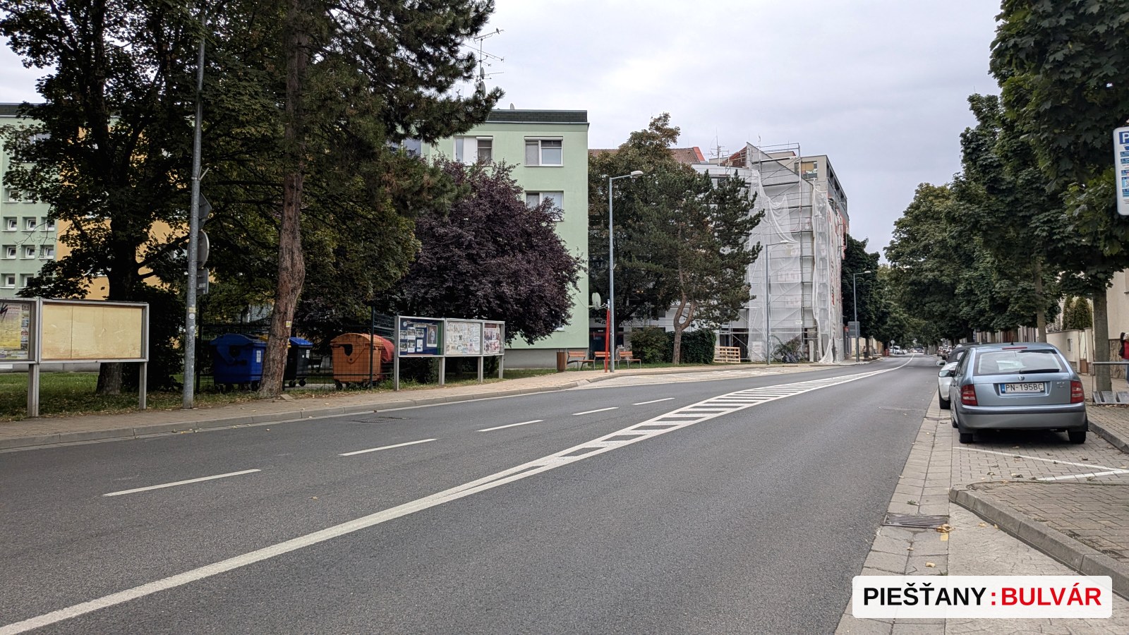 Fotografia ukazuje prázdnu mestskú ulicu s obytnými budovami. Na jednej z budov je viditeľné rusztovanie a ochranná sieť, čo znamená rekonštrukciu alebo výstavbu. Po oboch stranách sú zaparkované autá a niekoľko stromov s plnou zelenou korunou. Na ceste nie je žiadna snehová pokrývka ani jesenné listy. Obloha je oblačná.