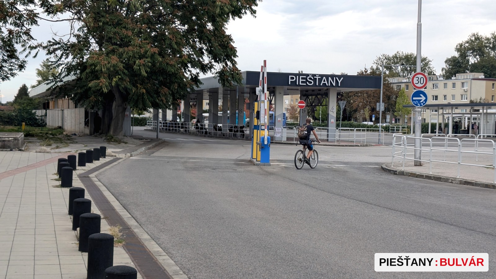 Redakčná fotografia vstupnej brány s nápisom Piešťany, cez ktorú prechádza cyklista. Okolie tvorí asfaltová cesta, chodník s čiernymi stĺpikmi a stromy. Na bráne je viditeľný veľký názov mesta.