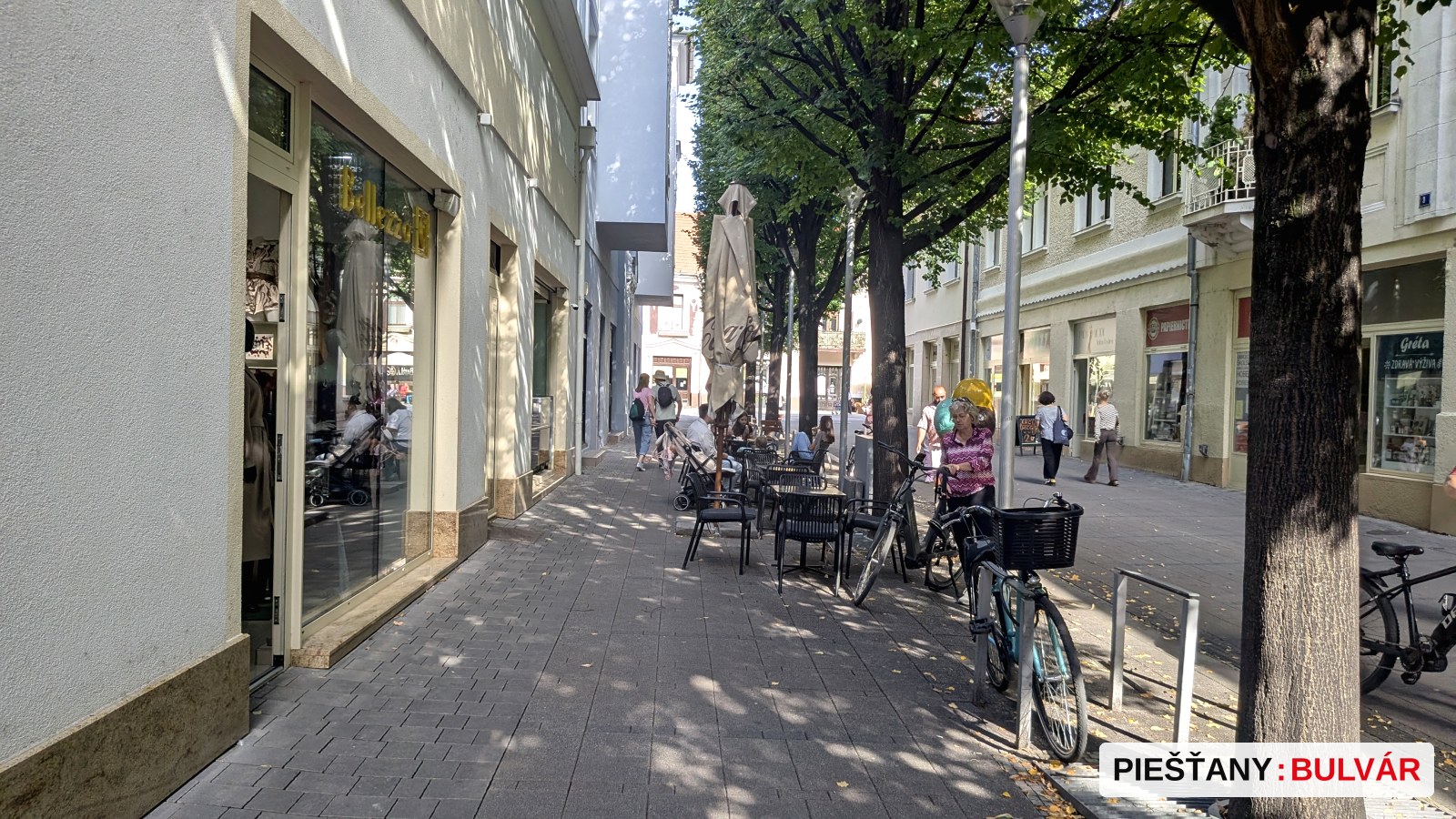 Mestská pešia zóna s kamenným chodníkom, lavičkami, posteľami s kaviarňou a stojanmi na bicykle. Niekoľko ľudí sa prechádza, slnečné počasie, stromy lemujú uličku.
