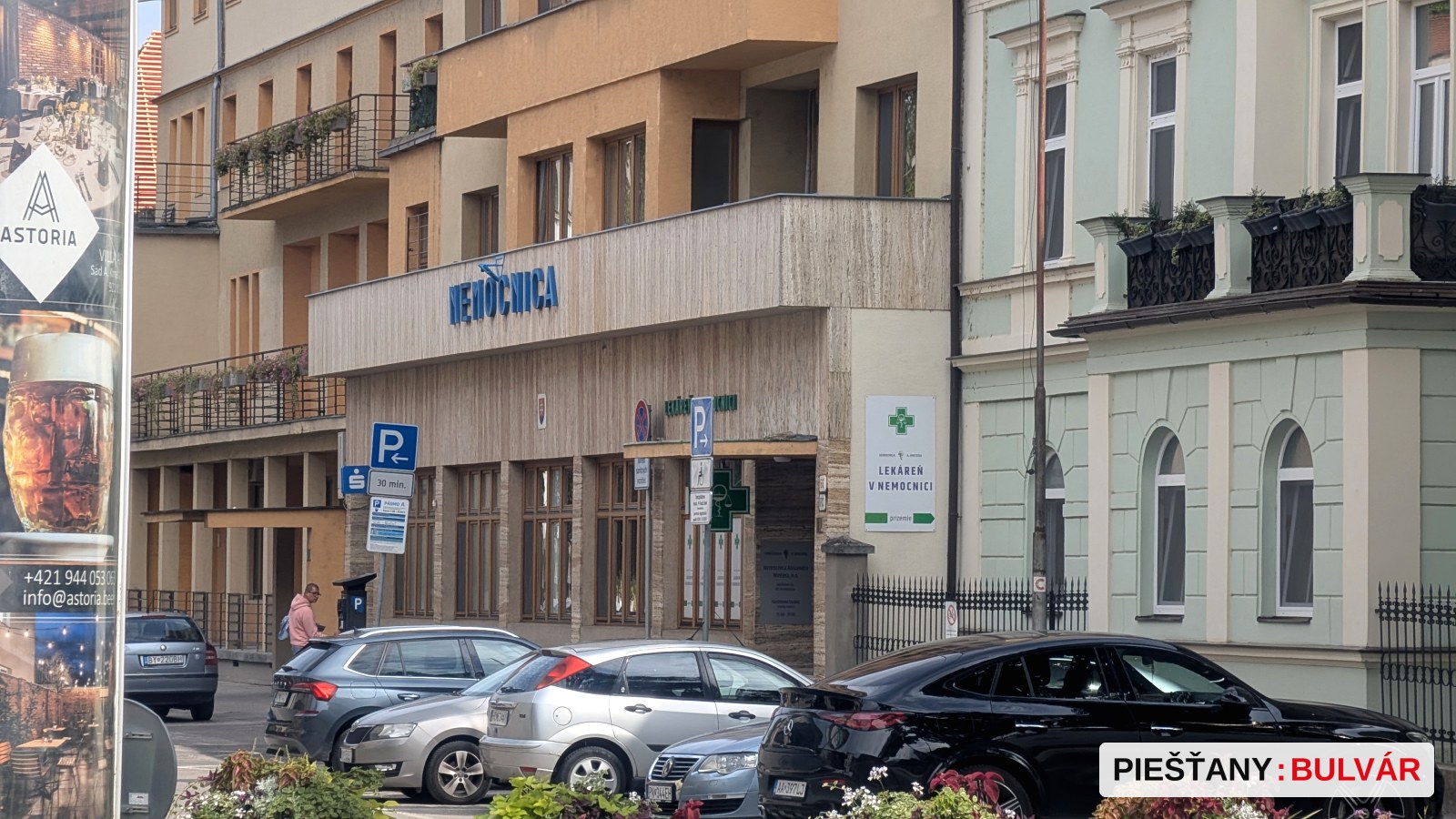 Fotografia zachytáva ulicu s parkovanými autami a budovou s nápisom Neúčtovnica. Vedľa budovy je viditeľný nápis Lekáreň Remedica. Na stĺpe pri ceste sú viaceré dopravné značky a parkovacie hodiny. Nevidno žiadne sezónne znaky ako sneh, kvitnutie či jesenné listy.