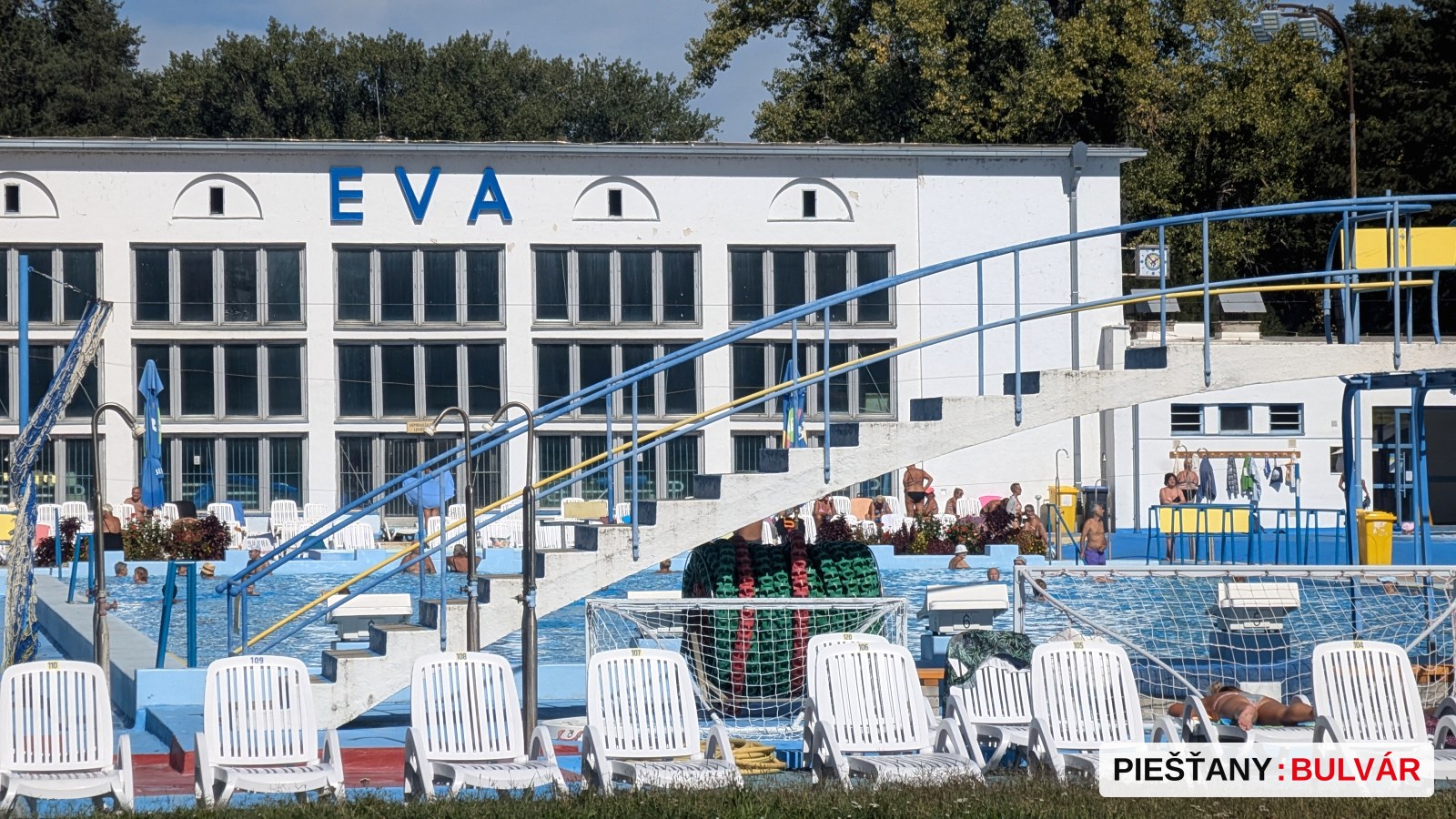 Fotografia ukazuje vonkajšie kúpalisko s bielymi plastovými lehátkami vpredu, nádrž plnú vody a šmýkačku alebo skokanskú dosku. Na pozadí je budova s veľkými oknami a nápisom EVA. Ľudia sú v bazéne aj okolo neho, počasie je jasné, slnečné.